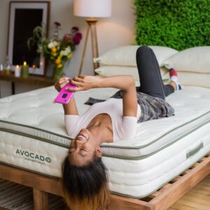 Woman resting her head all the way to the edge of the Avocado mattress demonstrating the superior edge support. She is laying on her back, reading her phone.